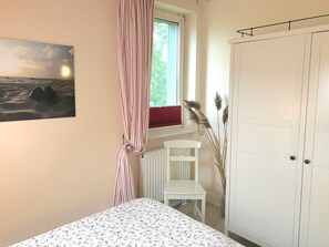 Een slaapkamer met een bed, een raam met gordijnen, een radiator, een witte stoel en een kledingkast.