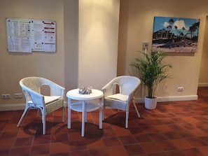 A room with two wicker chairs, a small round table with a decorative item, a potted plant, a bulletin board with notices, and a beach scene painting on the wall. - Ferienwohnung Achterdieck - Haus Borée (Prerow)