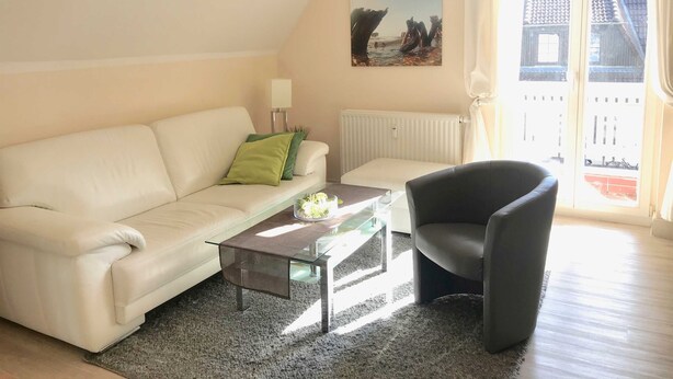 Ein Wohnzimmer mit einem weißen Sofa, einem schwarzen Sessel, einem Glastisch, einem Heizkörper und einem Bild an der Wand.