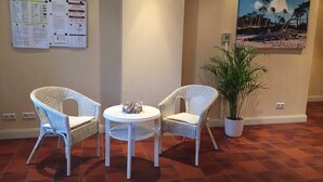 A room with two wicker chairs, a small round table with a decorative item, a potted plant, a bulletin board with notices, and a beach scene painting on the wall. - Vacation apartment Möövnest - House Borée (Prerow)