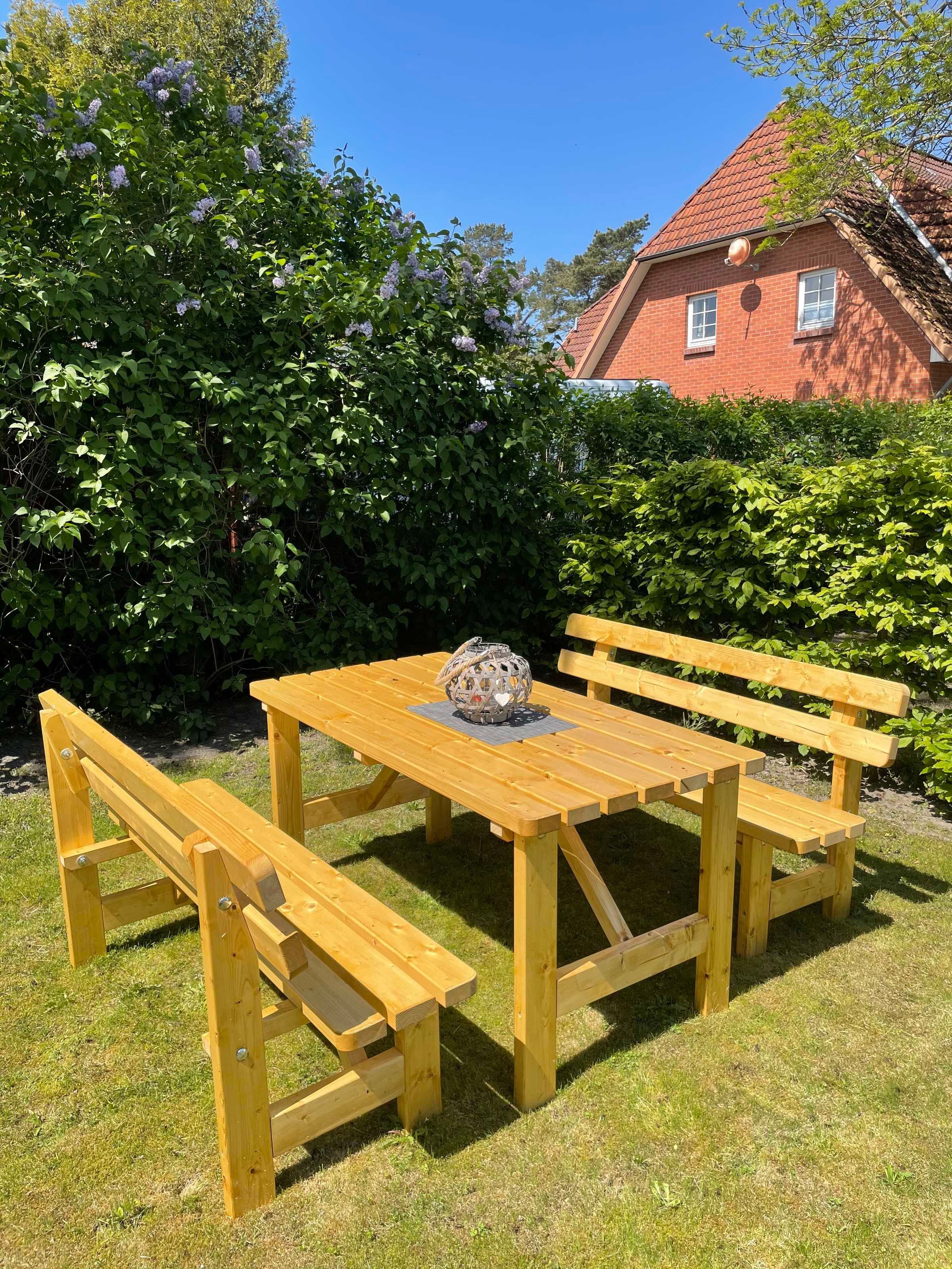 Ein hölzernes Picknicktisch mit Bänken, umgeben von üppigem Grün und einem roten Backsteinhaus im Hintergrund.