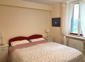 Une chambre à coucher avec un lit, des tables de chevet, une lampe, une fenêtre avec des rideaux et un tableau au mur.