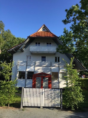 Un bâtiment à deux étages, doté d’un toit recouvert de tuiles rouges, d’un balcon à l’étage supérieur et d’une entrée fermée par un portail.