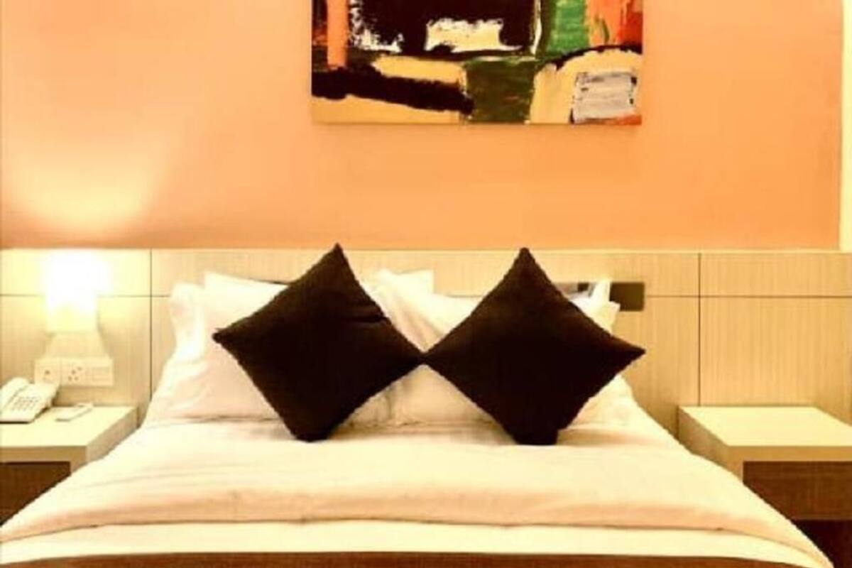 Hotellihuone, jossa on sänky, kaksi mustaa tyynyä, valkoinen päiväpeite, ruskea viltti, yöpöytä lampun kanssa ja taulu seinällä.