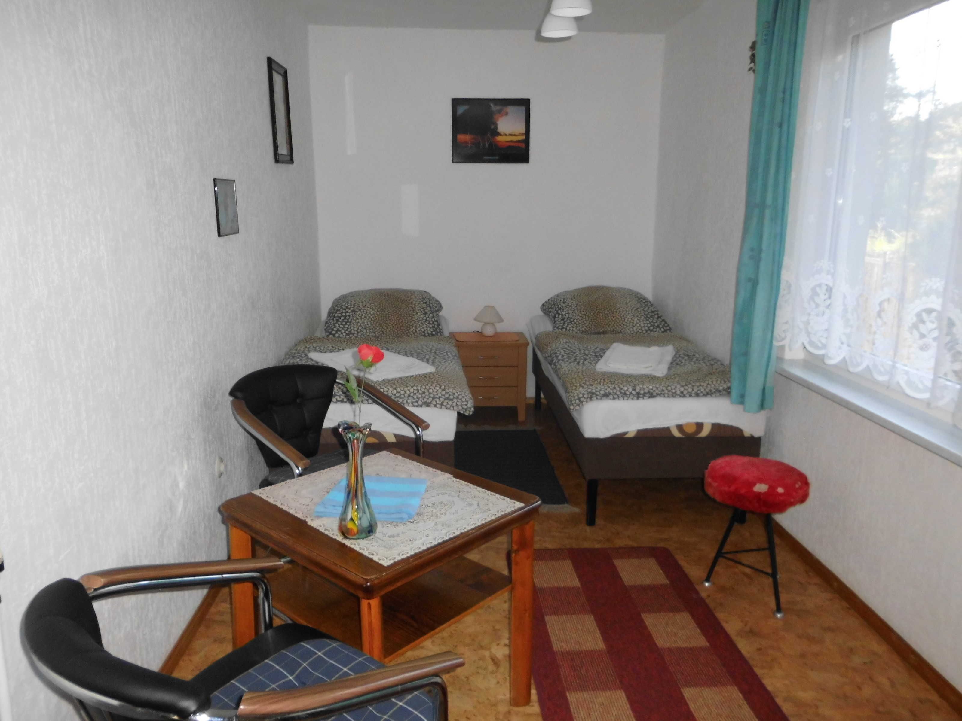 Una habitación pequeña y bien iluminada con dos camas, un escritorio, una silla y una ventana con cortinas.