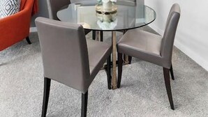 Et moderne spiseområde med et glasbord, stole, en dekorativ pendel og en grå væg med et indrammet spejl.