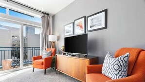 En moderne stue med en grå sofa, en orange lænestol og et tv-skab i træ. Der hænger tre billeder i rammer på væggen, et stort vindue med udsigt til bygninger, og en balkon med en plante.