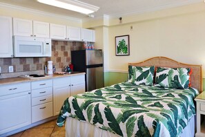 A compact kitchen with white cabinets, a microwave, and a refrigerator. There is a bed with a green leaf-patterned bedspread and pillows. - Ocean/City View Studio | Htd Pool |Balcony | Splash Park Next Door | Free Daily Activities Included (Daytona Beach)