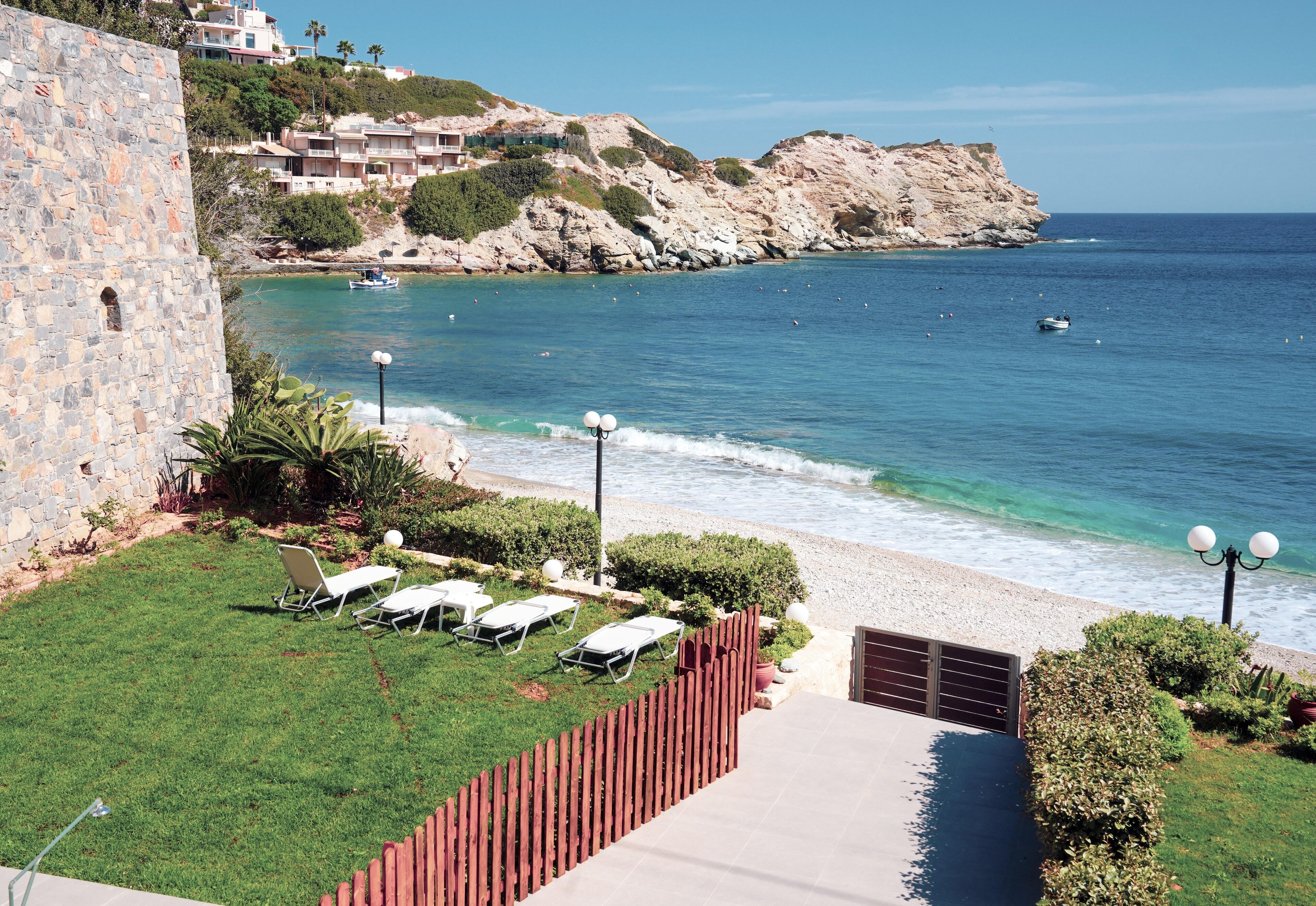 A coastal view with a stone wall, a grassy area with sun loungers, and a clear blue sea.