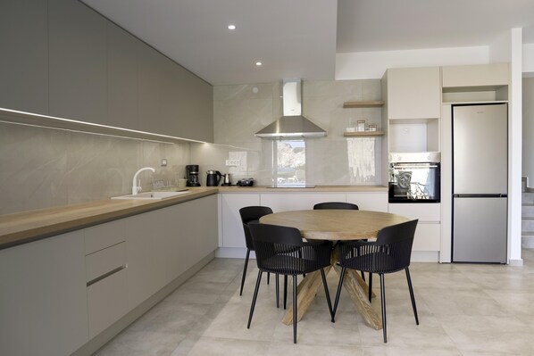 A modern kitchen with a wooden dining table and black chairs, a stainless steel range hood, and a built-in oven. - DreamWave Residence (Malevizi)