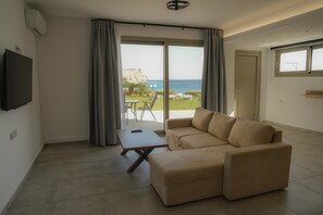 Una sala moderna con un sofá, una mesa de centro y un televisor. Hay un ventanal corredero que da a un espacio exterior con vista al mar.