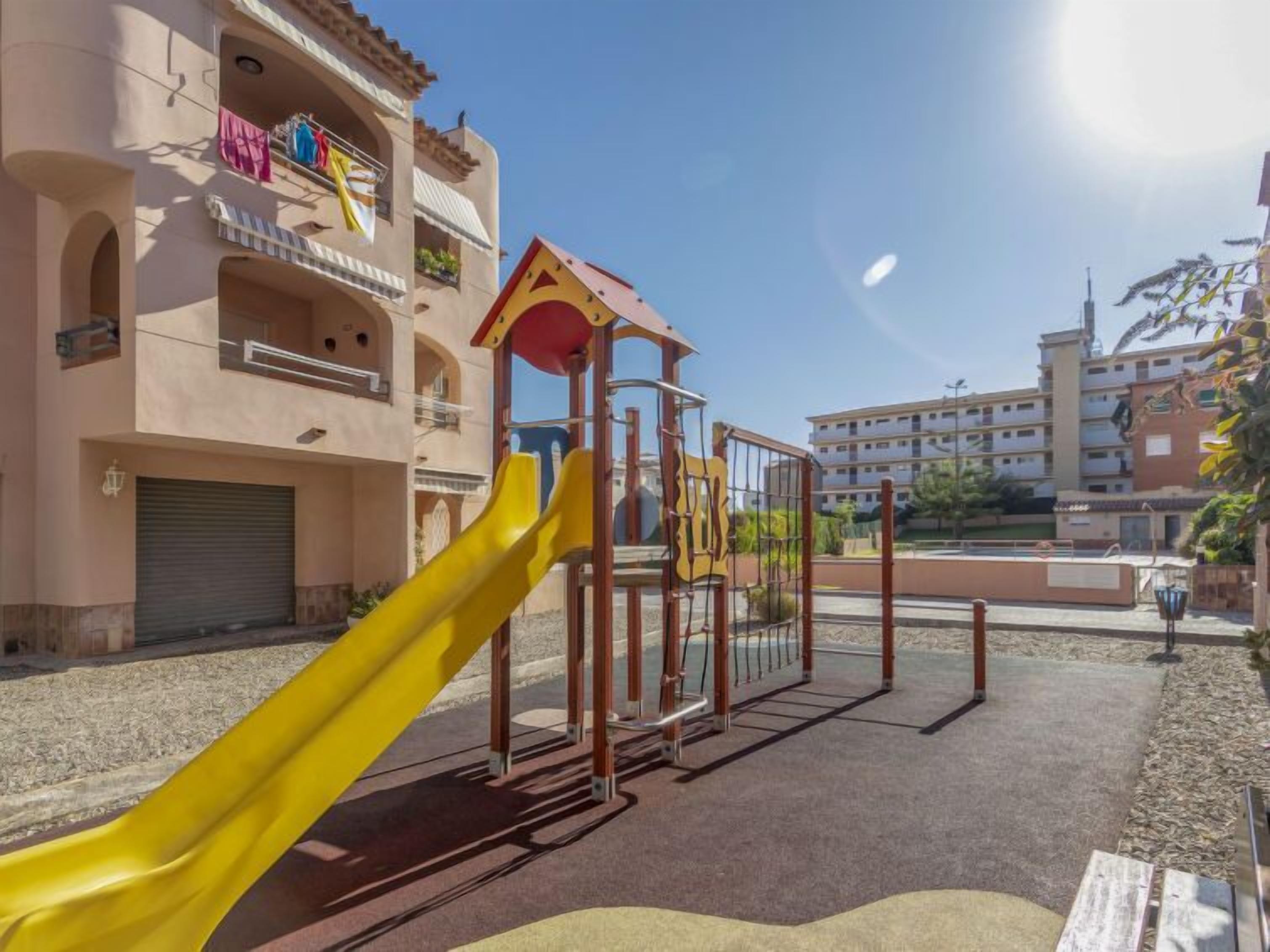 A playground with a yellow slide and a climbing structure, set against a building with balconies and a clear sky.