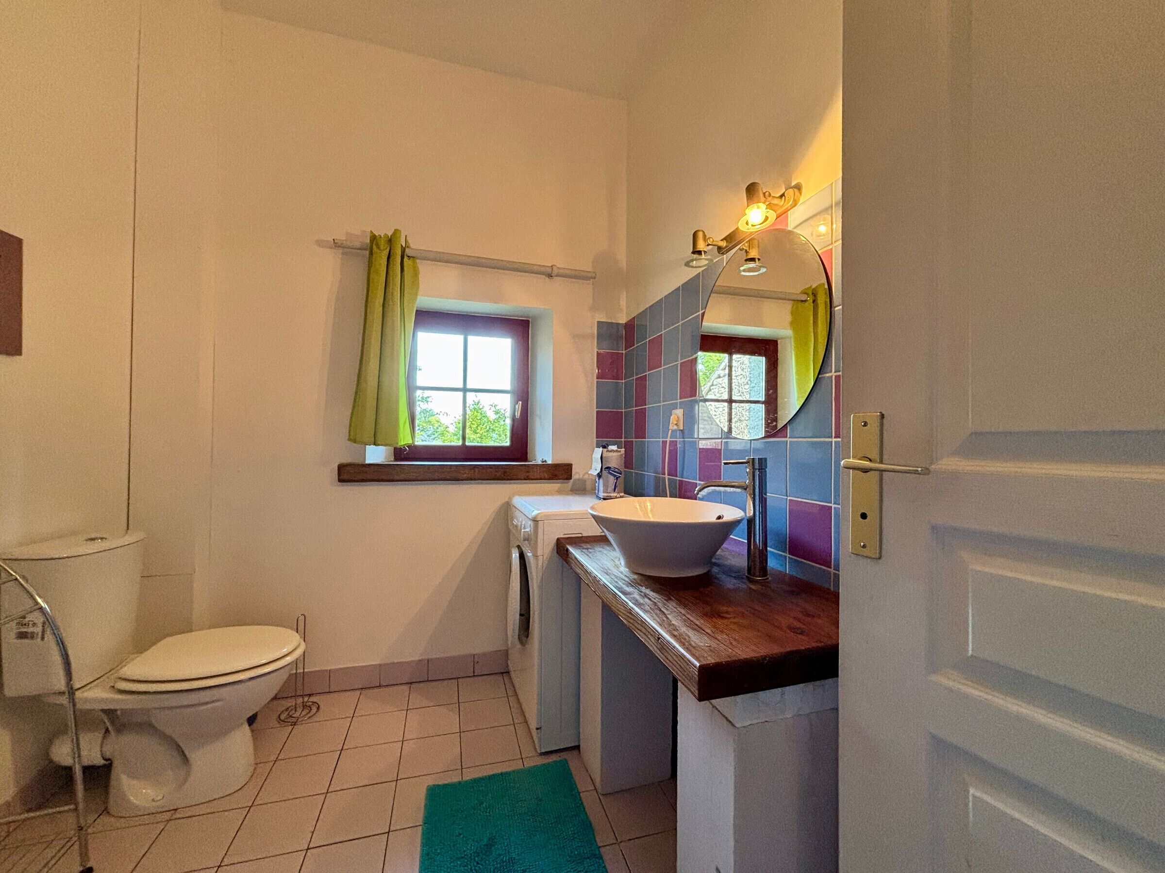 A compact bathroom with a toilet, washing machine, sink, and a window with curtains.
