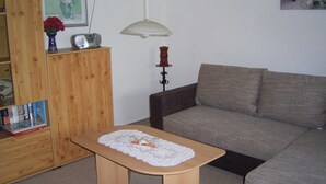 A living room with a wooden cabinet, a table with a lace tablecloth, a grey sofa, a hanging lamp, and a framed picture on the wall. - Vacation apartment in Seedorf - Holiday apartment Kontschack (Sellin)