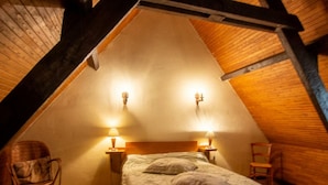A cozy attic bedroom with a sloped ceiling, two bedside lamps, a bed with a quilt, a chair, and a small rug.