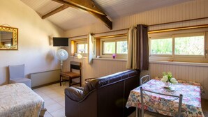 A cozy room with a sofa, a small table with a floral tablecloth, a chair, a bed, a TV, and windows with curtains. - Charming studio near Sarlat for a peaceful getaway (La Roque-Gageac)