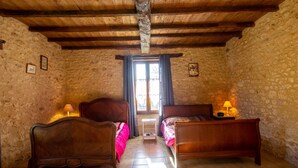 Une pièce rustique aux murs de pierre, avec des poutres en bois, deux lits, une fenêtre avec des rideaux et une petite table sur laquelle se trouve une lampe.