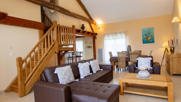 Amplia sala de estar con escalera de madera, sofás de cuero, zona de comedor y vistas al exterior.