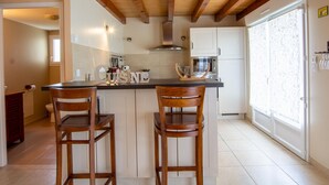 Una cocina con vigas de madera, una isla central con dos taburetes de bar, armarios blancos y un ventanal con cortinas estampadas.