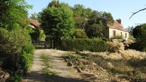 Ein ländlicher Pfad führt zu einem Steinhaus mit rotem Dach, umgeben von üppiger Vegetation und einem Holzzaun.