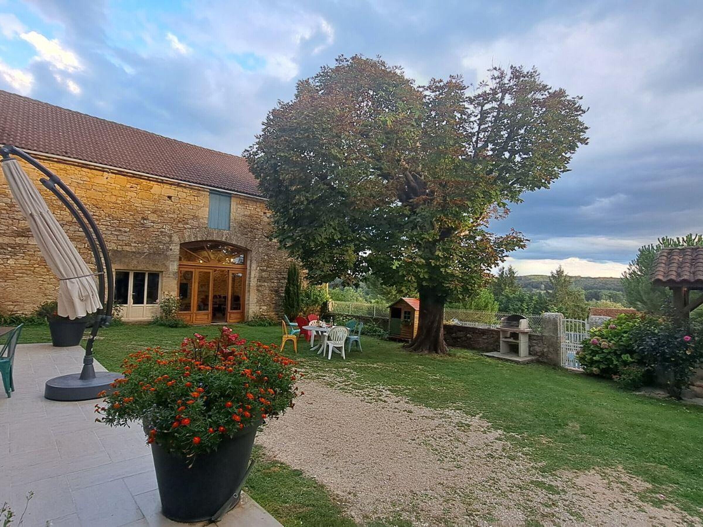 A stone building with a tiled roof, a large tree, and outdoor seating.