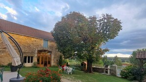 A stone building with a tiled roof, a large tree, and outdoor seating.