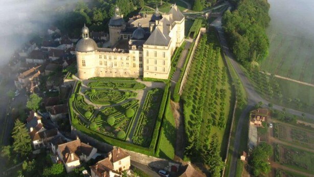 An aerial view of a historic estate with a large central building, formal gardens, and a surrounding forested area.