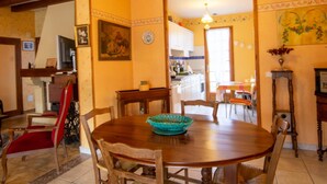 A dining area with a wooden table and chairs, a chandelier, and a kitchen in the background.