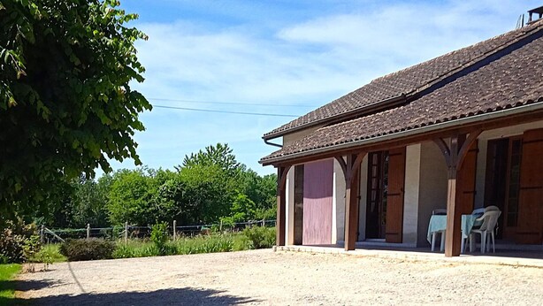 A building with a covered porch, a gravel driveway, and a fenced area with greenery.