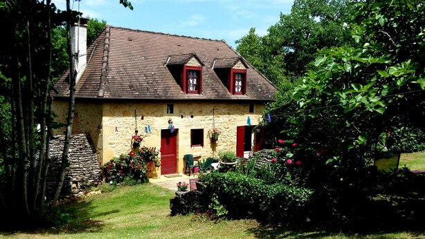 Una casa de piedra con techo inclinado, dos puertas rojas y un pequeño jardín con flores y césped.