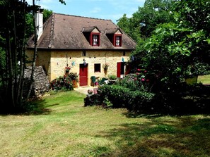 A stone house with a sloped roof, two red doors, and a small garden area with flowers and a lawn. - Charming house in Dordogne with garden and fireplace (Sainte-Mondane)