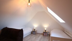 A small, well-lit attic bedroom with a sloped ceiling, two beds, a chair, and a skylight.