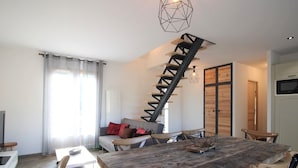 A modern living room with a wooden dining table, a sofa with cushions, a staircase, and a kitchen area.