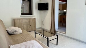 A modern living room with a sofa, a glass coffee table, a TV mounted on the wall, and a sliding door leading to a kitchen area.