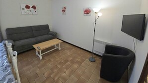 A small, furnished room with a grey sofa, a wooden table, a floor lamp, a black chair, and a mounted TV. - Charming house near Domme with Wifi, 2 bedrooms (Domme)