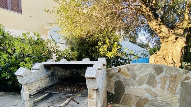 Una barbacoa de pedra exterior amb una reixa metàl·lica, envoltada de vegetació i un arbre.