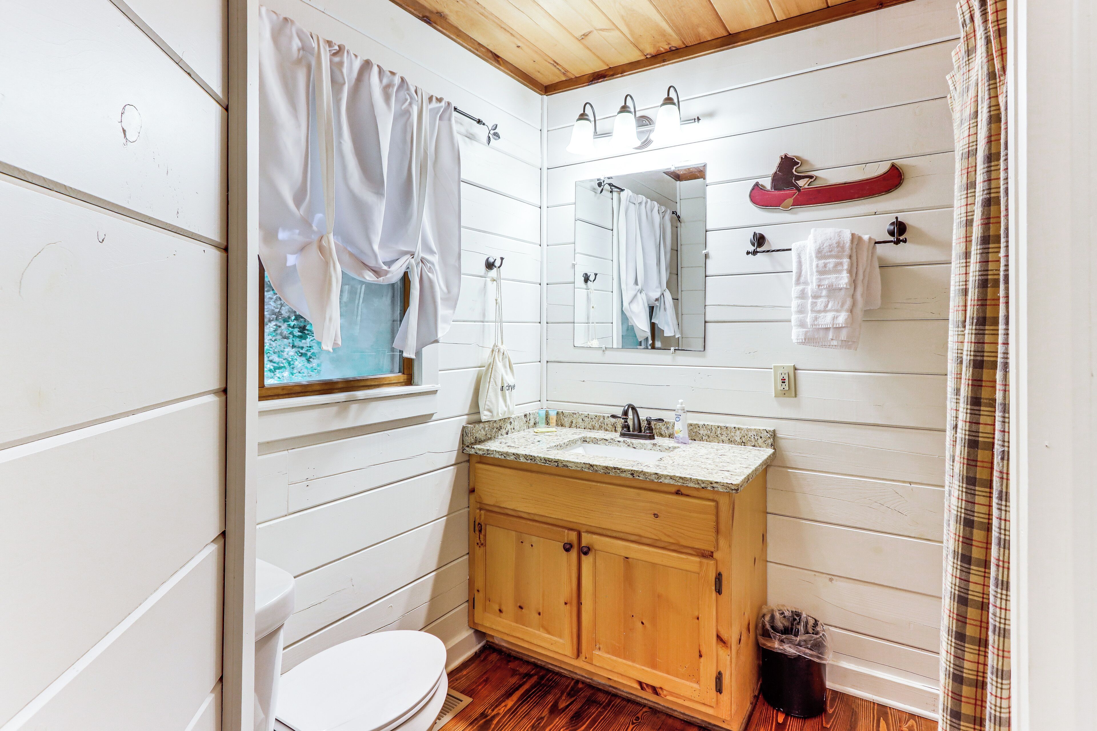 A bathroom with a wooden vanity, a white toilet, a window with a view of trees, and a mirror with a canoe decoration.