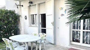 A white outdoor dining area with a round table and four chairs, a door with a window, and a small plant to the left. - M282 - Marcelli, three-room apartment with garden 100m from the sea (Marcelli)