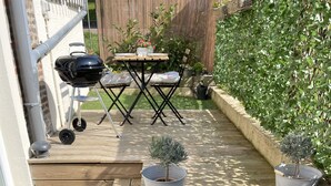 Eine Holzterrasse mit Tisch und Stühlen, umgeben von Kübelpflanzen und einem Grill.