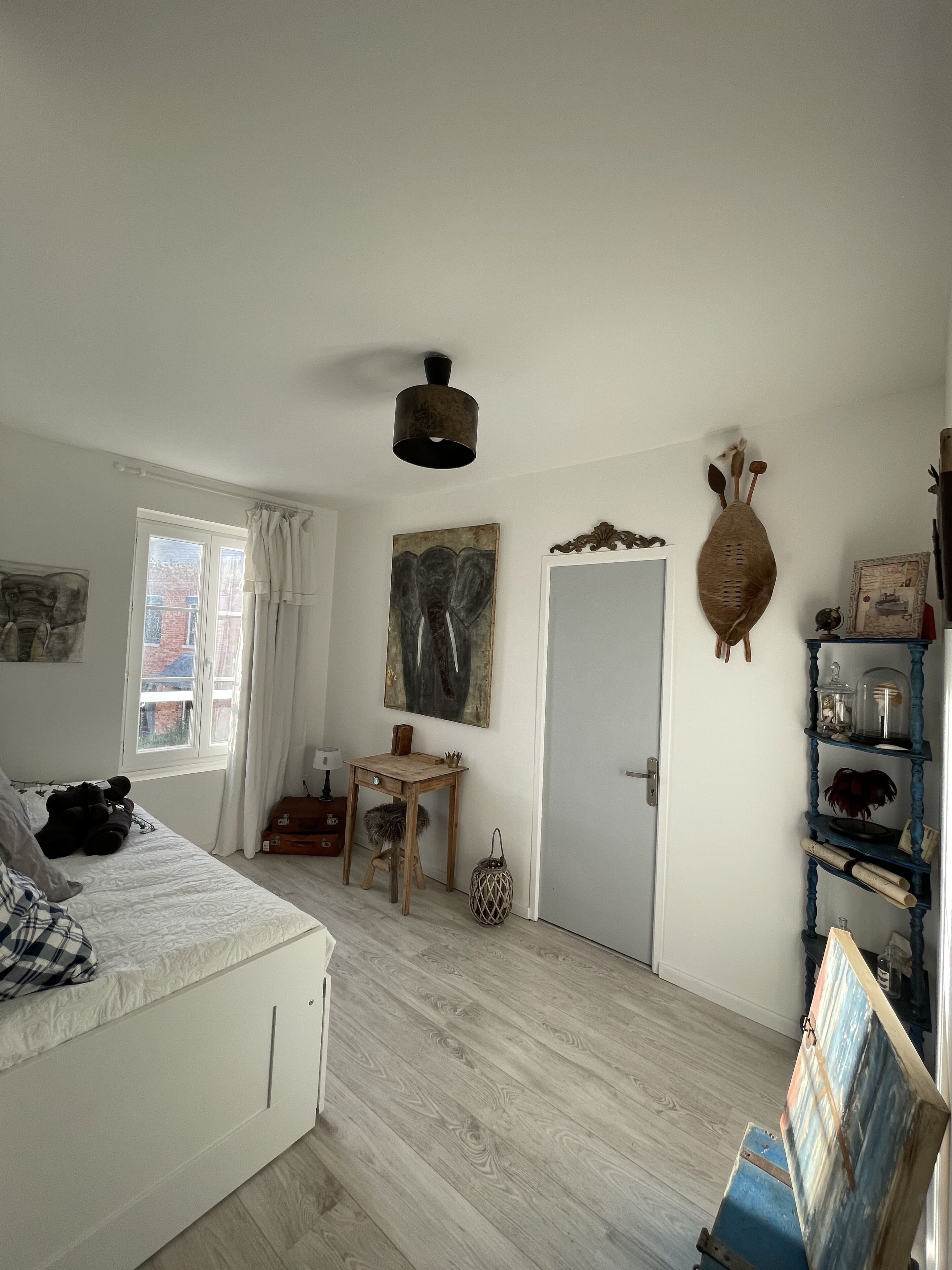 A bedroom with a bed, a desk, a chair, a lamp, a painting, a door, and a shelf with decorative items.