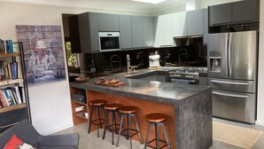 A modern kitchen with a central island, bar stools, stainless steel appliances, and a bookshelf with books. - thespaceperth- the funkiest bali style villa in Perth (Maida Vale)