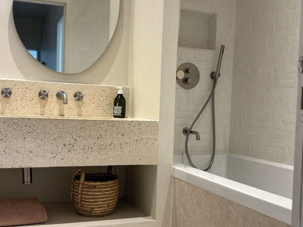A bathroom with a granite countertop, a round mirror, a shower with a handheld showerhead, and a bottle of soap.