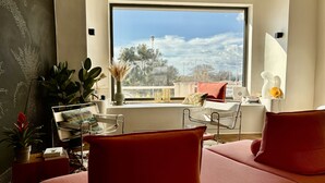 A modern living room with a large window, a sofa, a side table, and a potted plant. - House facing the sea, bright, air-conditioned, terrace, family, calanques (Marseille)