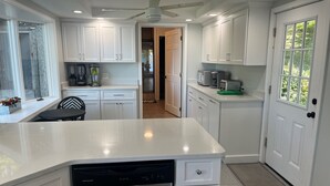 A modern kitchen with white cabinetry, a dishwasher, a ceiling fan, and a window with a view of greenery.