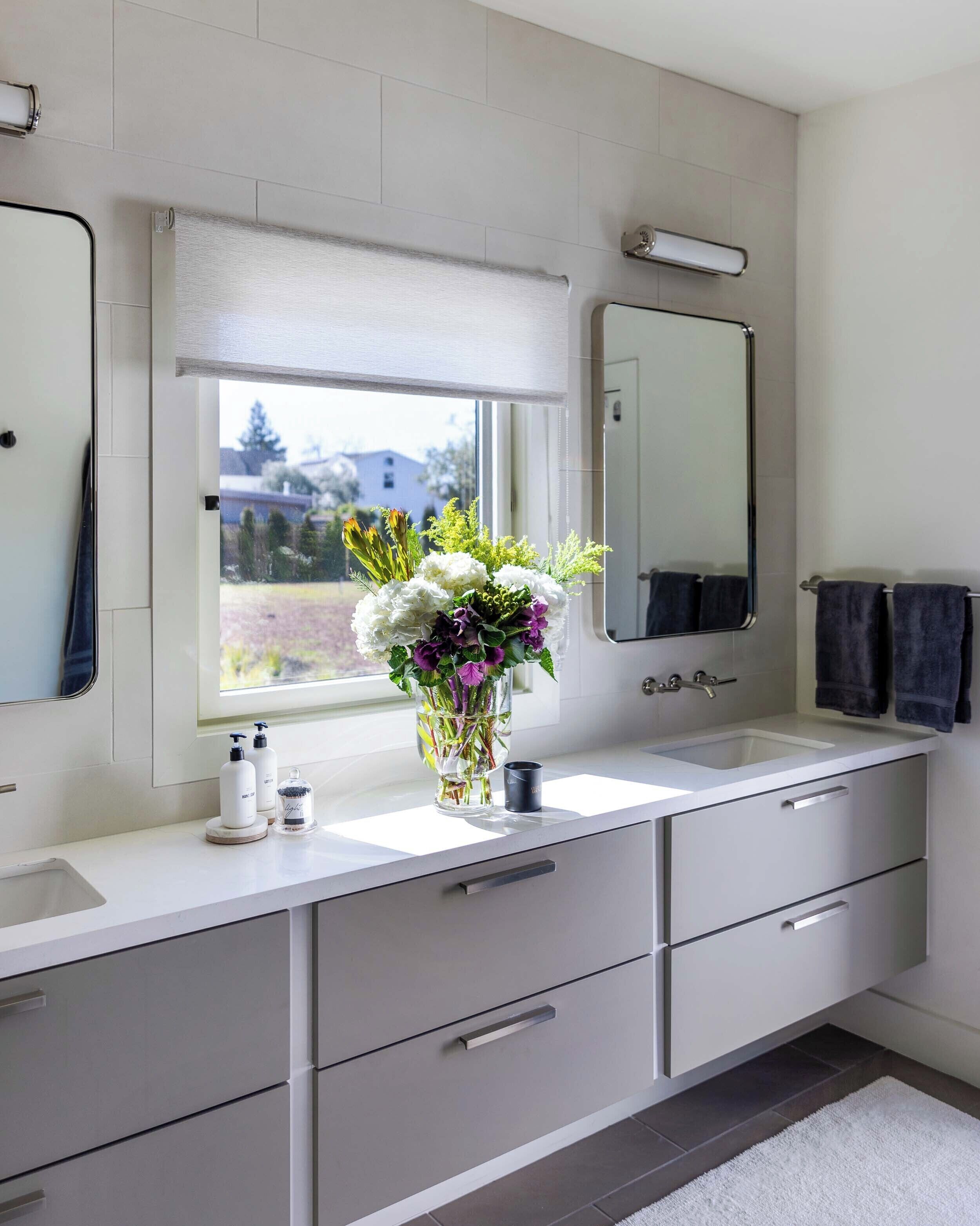 A modern bathroom with two sinks, a large mirror, and a window with a view of greenery.