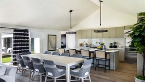 A modern kitchen with a dining area, featuring a large island, pendant lighting, and a potted plant.