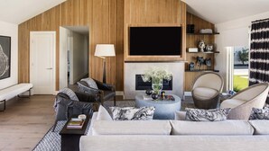 A modern living room with a high ceiling, a fireplace, a flat-screen TV, and a seating area with a sofa and chairs.