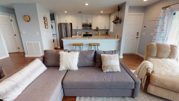 Una sala moderna con un sofá seccional gris, dos taburetes de bar y un sillón color beige. Se ve una cocina al fondo con gabinetes blancos y electrodomésticos de acero inoxidable.