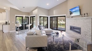 A spacious living room with a fireplace, large windows, and a modern kitchen in the background. - Stunning Home | Pool, Dock & Direct Gulf Access (Marco Island)