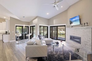 A spacious living room with a fireplace, large windows, and a modern kitchen in the background. - Stunning Home | Pool, Dock & Direct Gulf Access (Marco Island)
